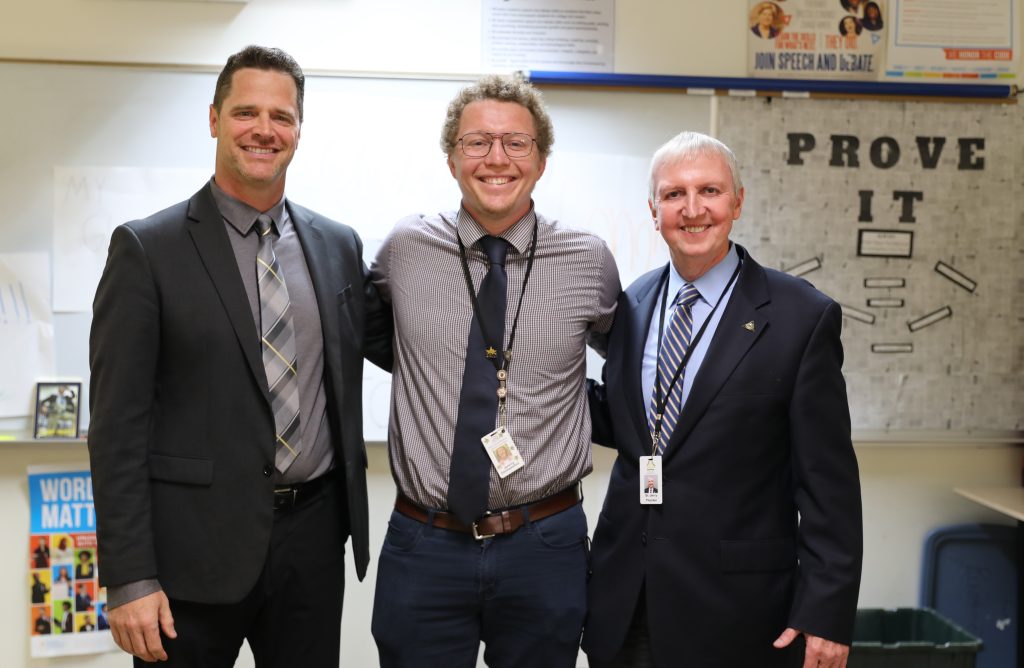 Three men posing together in a room, one of them wearing a tie. The man on the left is smiling and has his arm around the